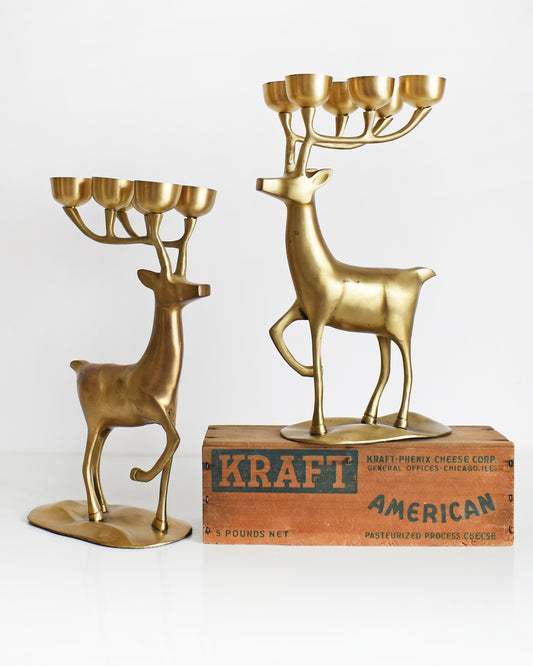 A pair of large vintage brass deer candelabras that have six tea light candle holders on top of their antlers. One of the deer is standing on a brown box.