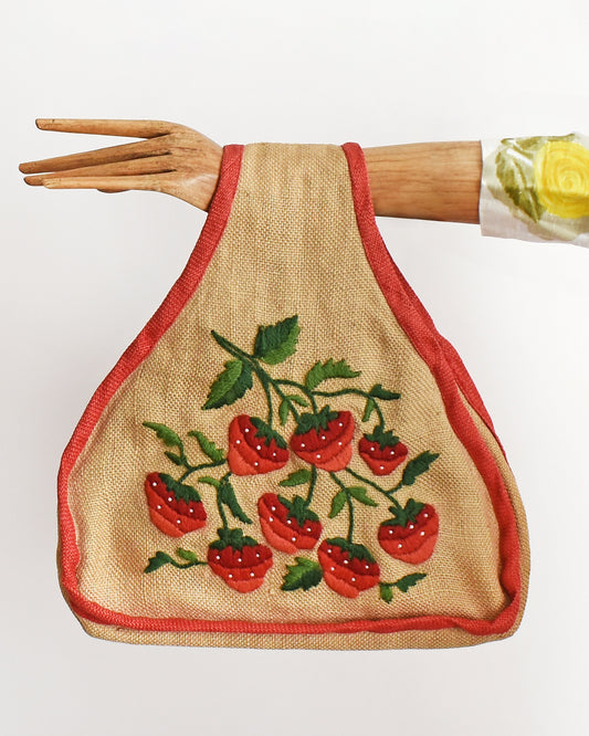 A vintage 60s burlap purse that has  embroidered strawberries on the front, with green leaves and small white pearl beaded seeds and red trim. The purse is being modeled by a wooden arm