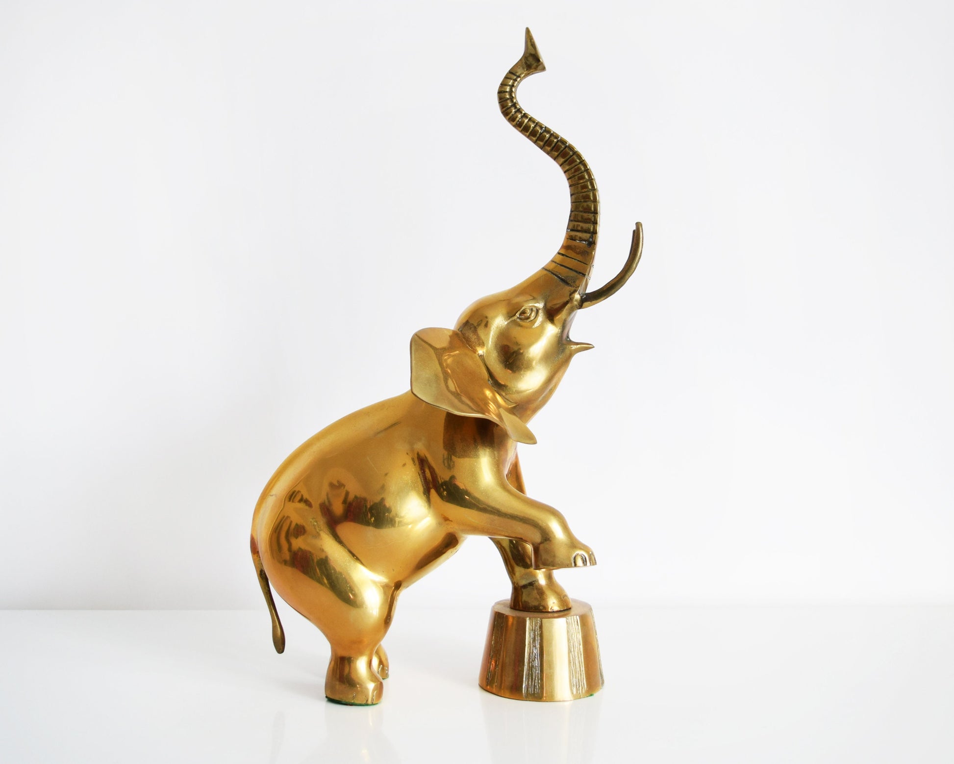 Side view of a large brass elephant that is balancing one of its front legs on a a platform and proudly displaying its trunk up for good luck.