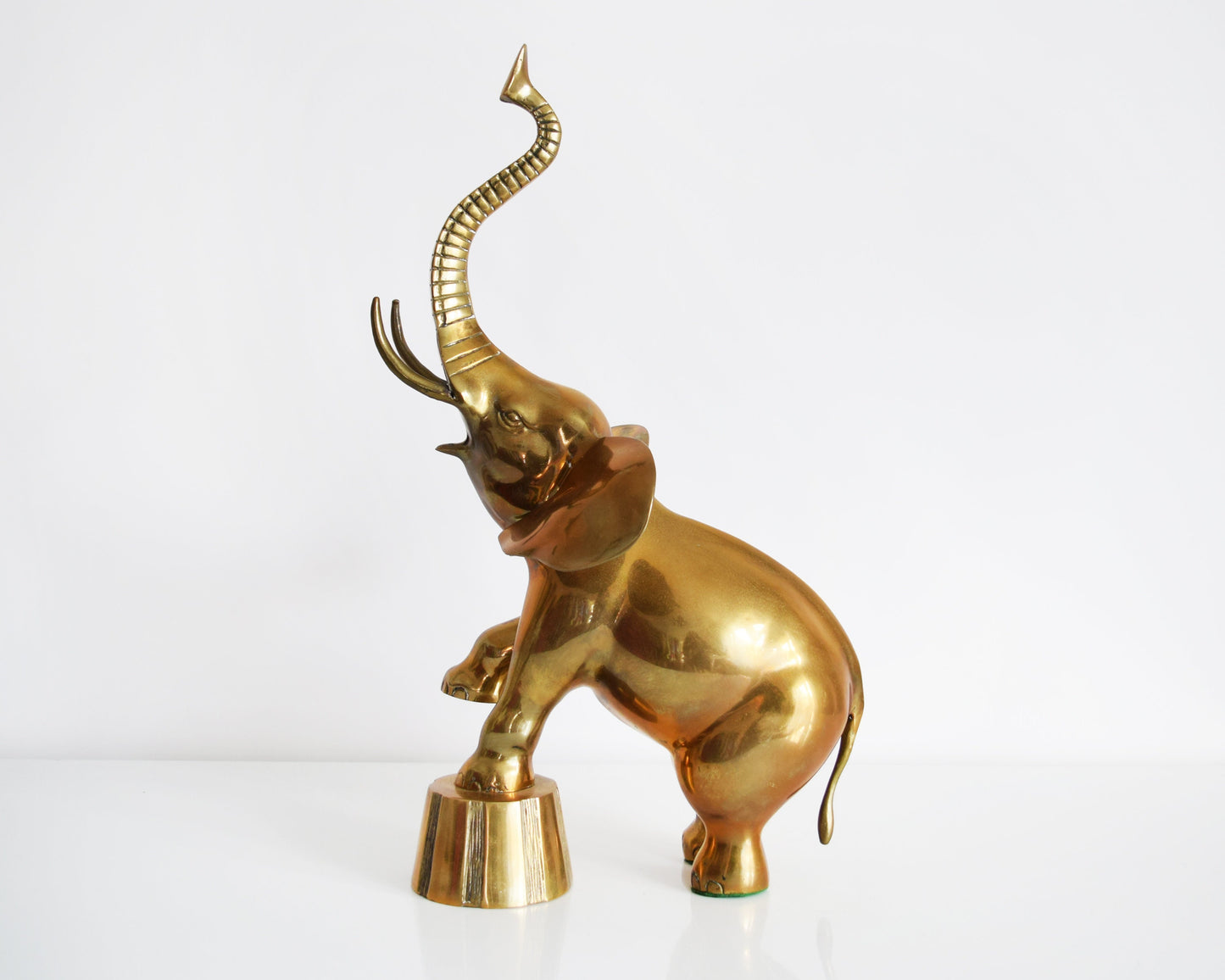 Side view of a large brass elephant that is balancing one of its front legs on a a platform and proudly displaying its trunk up for good luck.