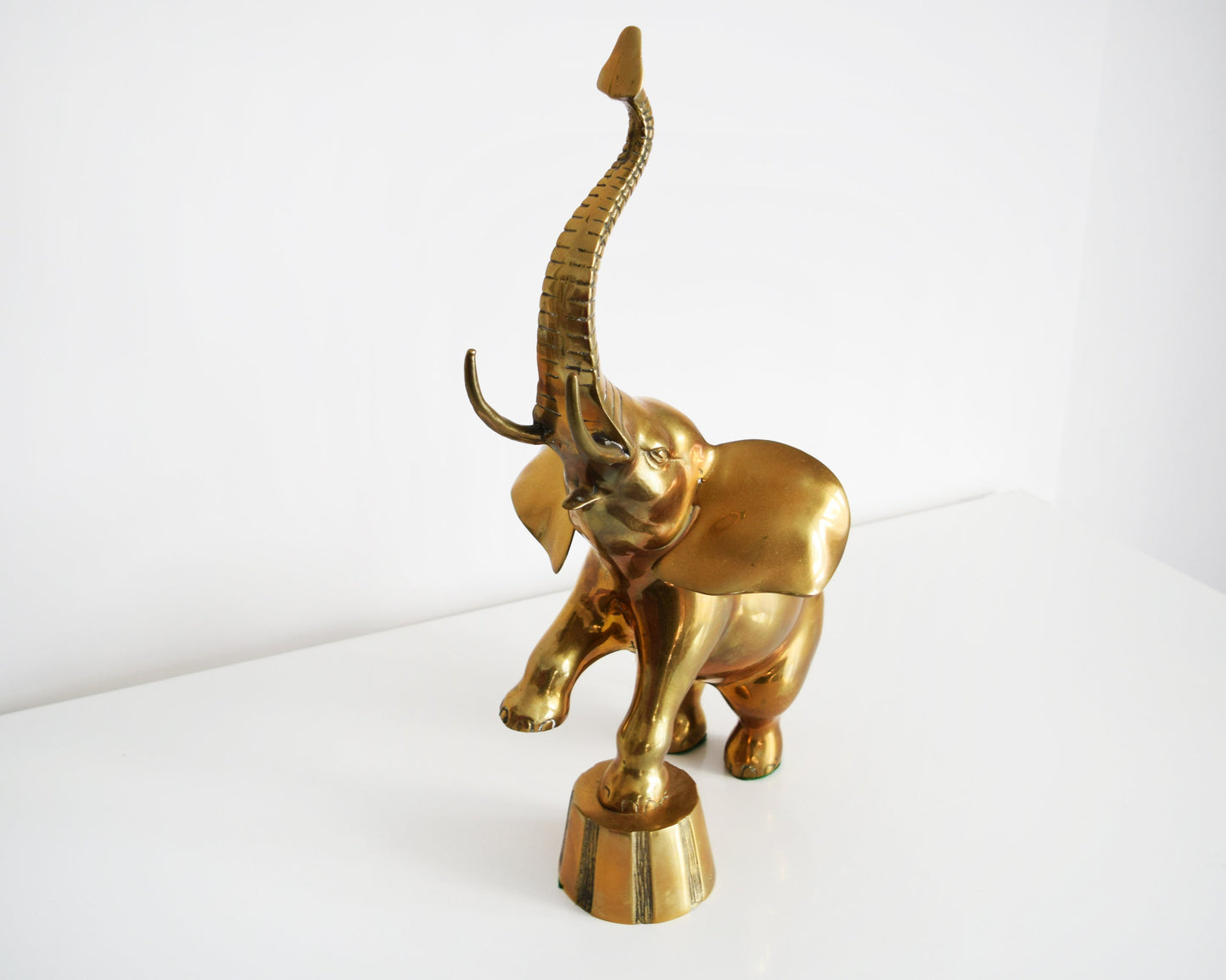 Side upper view of a large brass elephant that is balancing one of its front legs on a a platform and proudly displaying its trunk up for good luck.