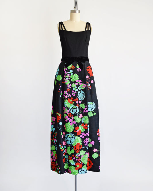 A vintage 60s black maxi dress that has a colorful floral print on the skirt and a black velvet bow at the waist.