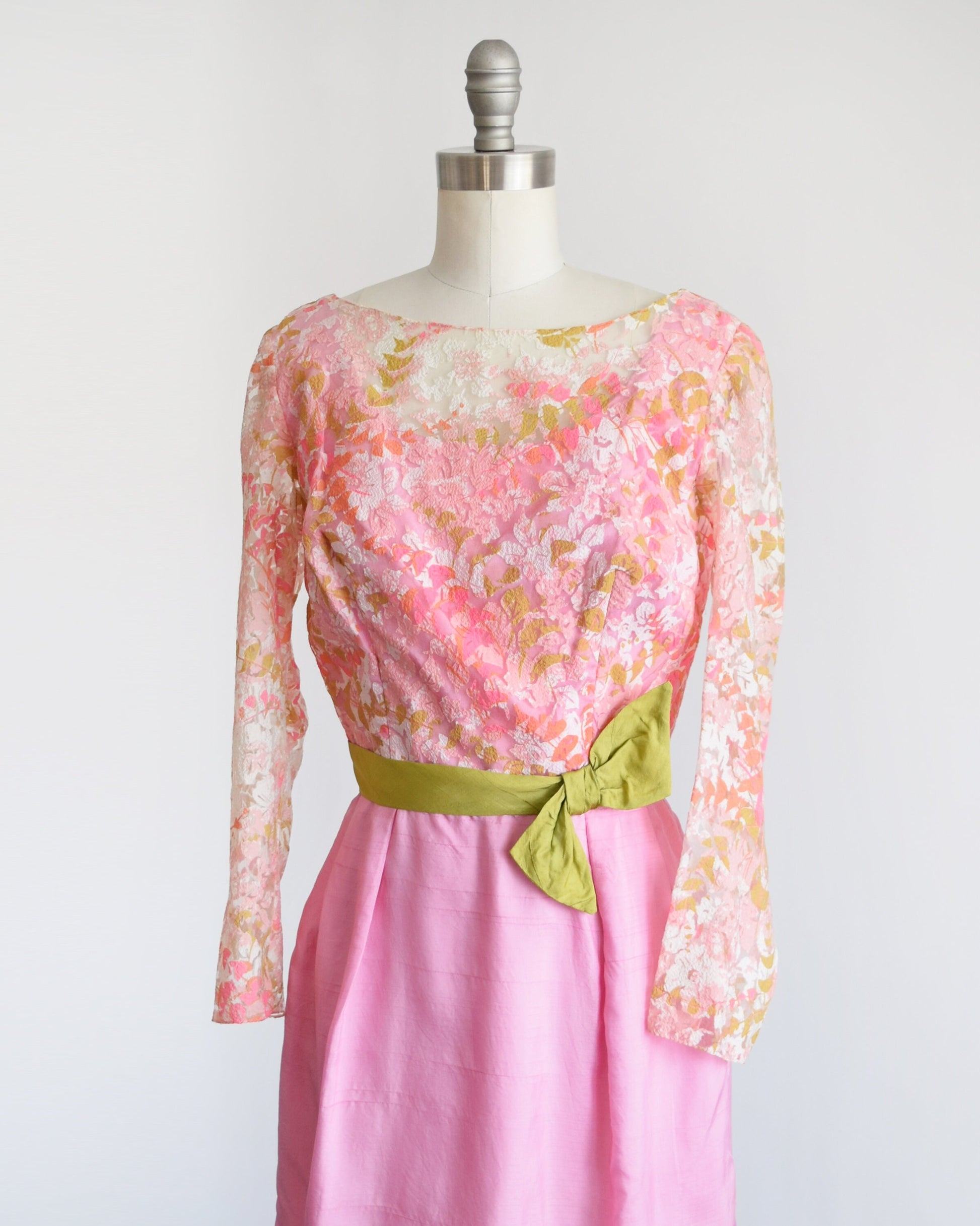 Side front view of a vintage 60s dress that has a pink, green, and white sheer floral lace bodice. Long sleeves with small slits on the inside of the cuffs. Fitted waist that's adorned with a green bow sash belt which hooks on the back. 
