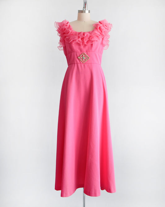 A vintage 1970s hot pink maxi dress that has a ruffled neckline and a gold tone rhinestone brooch at the waist.