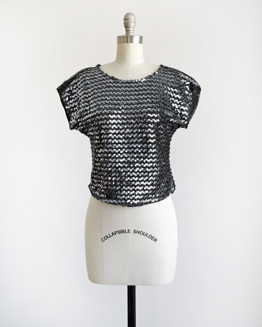 A vintage 80s sequin top that has silvery sequins set on the front on a metallic silver and black background. Matching black and silver lurex back. Cap sleeves. Rounded waistline. 