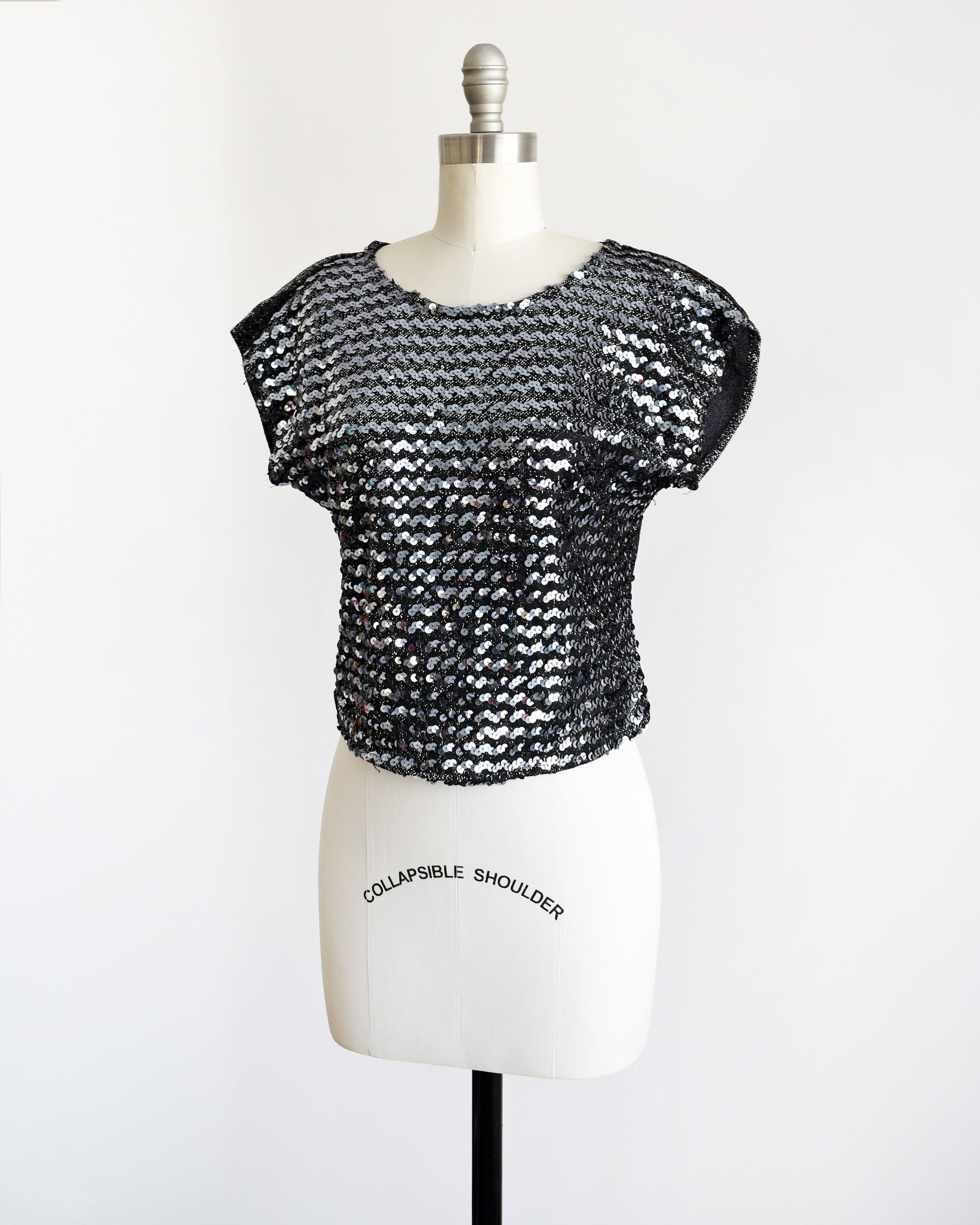 Side front view of a vintage 80s sequin top that has silvery sequins set on the front on a metallic silver and black background. Matching black and silver lurex back. Cap sleeves. Rounded waistline. 