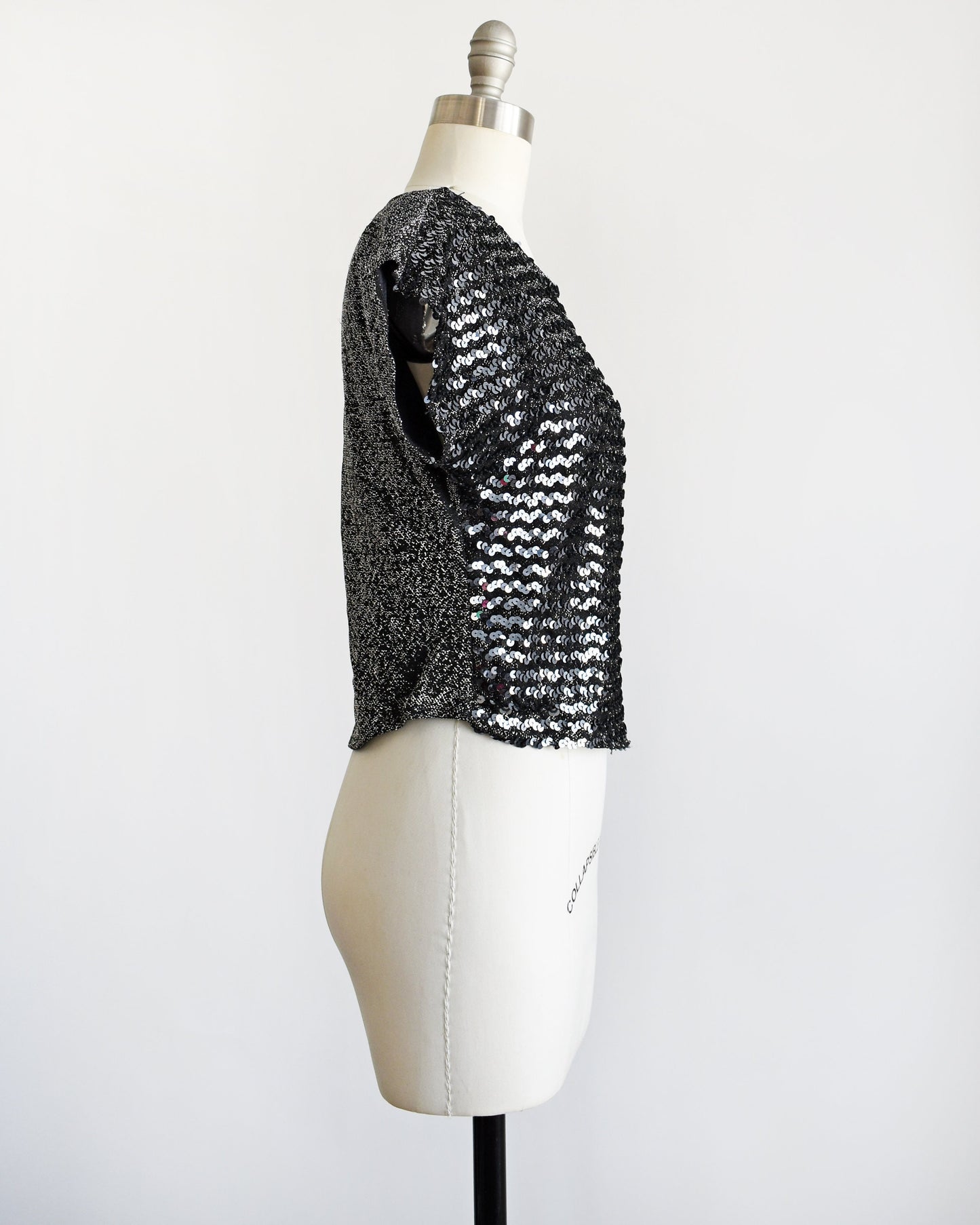 Side view of a vintage 80s sequin top that has silvery sequins set on the front on a metallic silver and black background. Matching black and silver lurex back. Cap sleeves. Rounded waistline. 