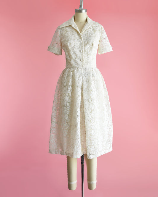 A vintage cream floral lace dress with collared neckline, short sleeves, and button front on a dress form set on a pink background