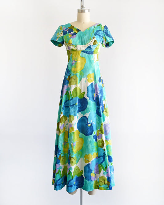 A vintage Hawaiian maxi dress on a dress form. The dress is covered with blue and green flowers that looks like a watercolor print. The dress has short sleeves, an empire waist, and a long skirt.