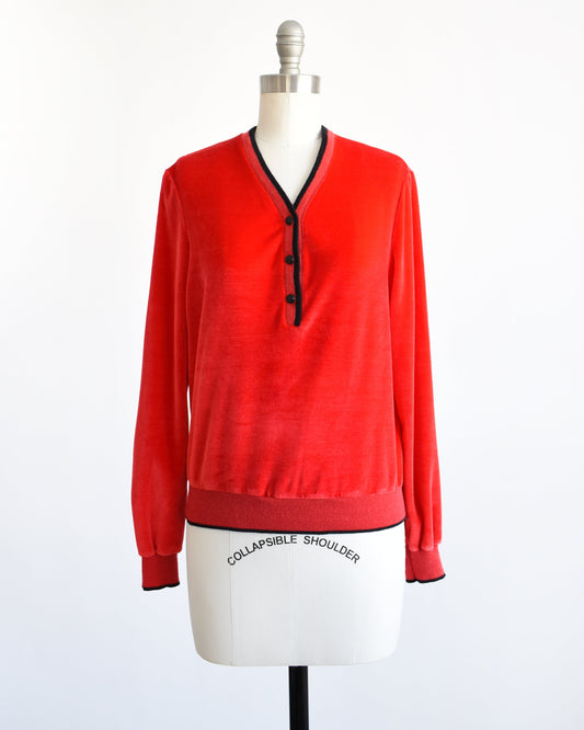 A vintage 70s red velour top with black trim along the v-neckline, cuffs, and hem. There are three black buttons on the front and the top is modeled on a dress form.