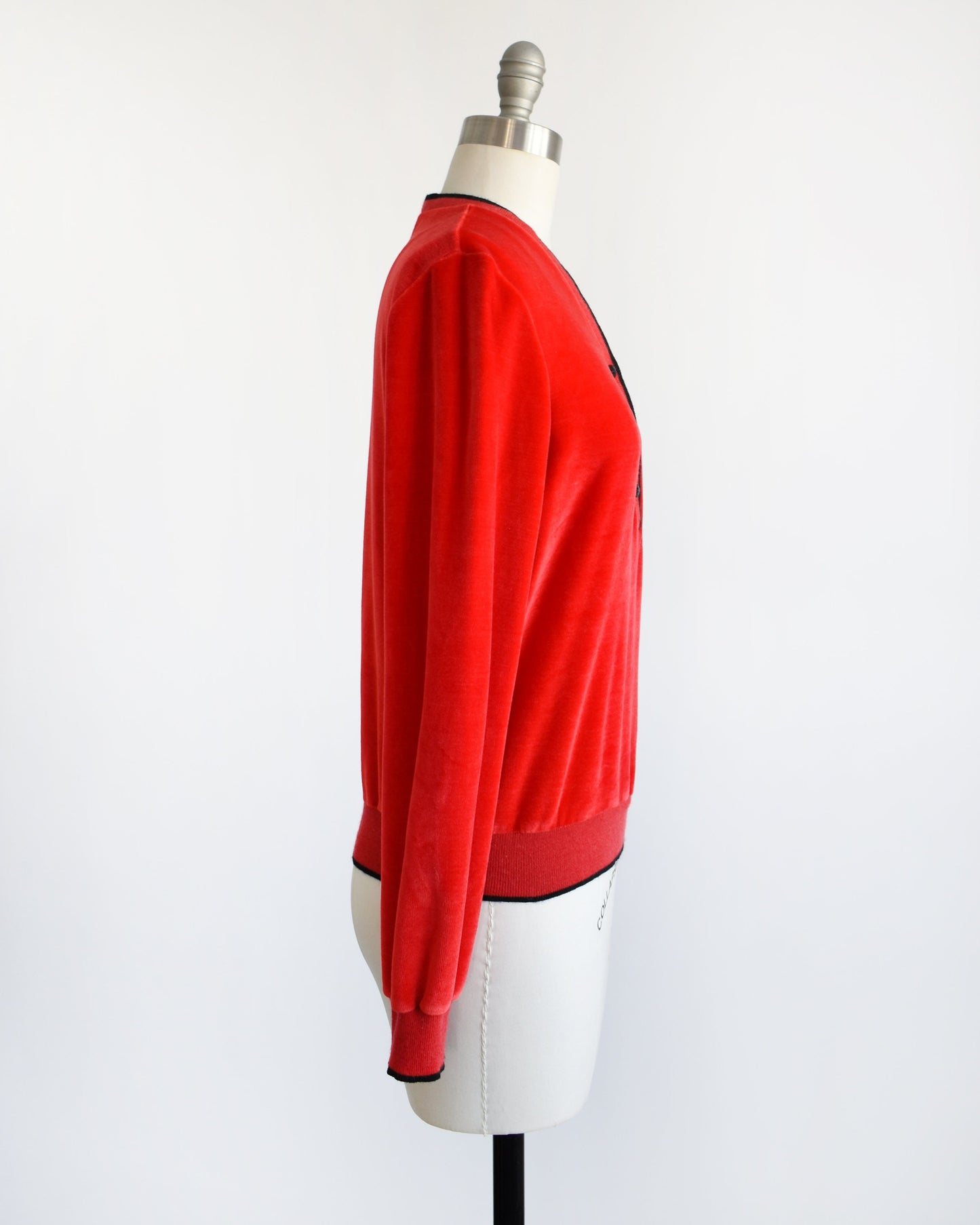 Side view of a vintage 70s red velour top with black trim along the v-neckline, cuffs, and hem. There are three black buttons on the front and the top is modeled on a dress form.