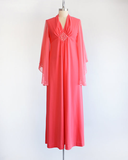 A vintage 70s coral pink maxi dress that has a v-neckline, rhinestones on the waist, and semi-sheer long angel sleeves with matching rhinestone trim. The dress is modeled on a dress form.