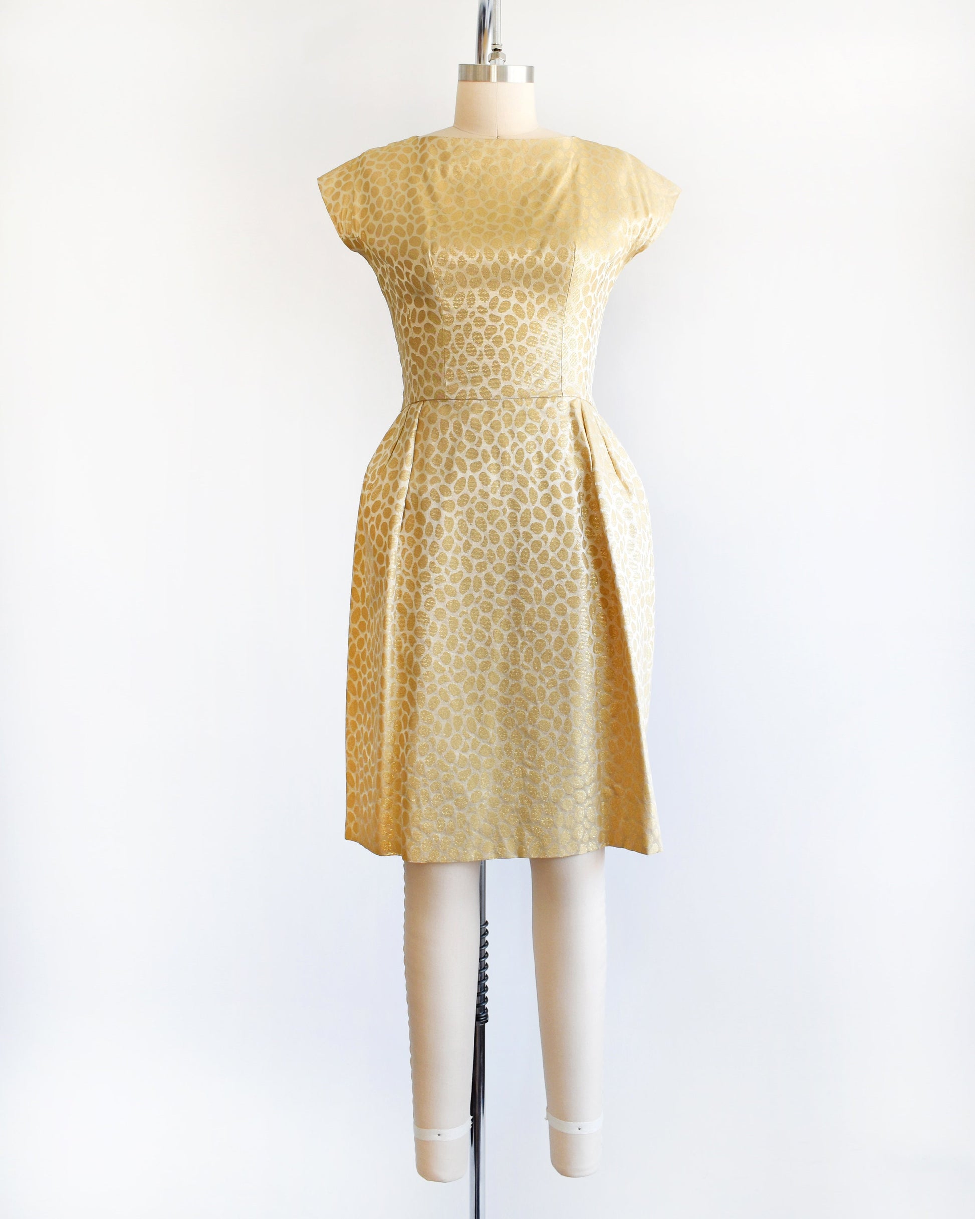 A vintage 60s gold dress with metallic gold brocade polka dots. The dress features two large pleats on the front of the skirt. The dress is modeled on a dress form.