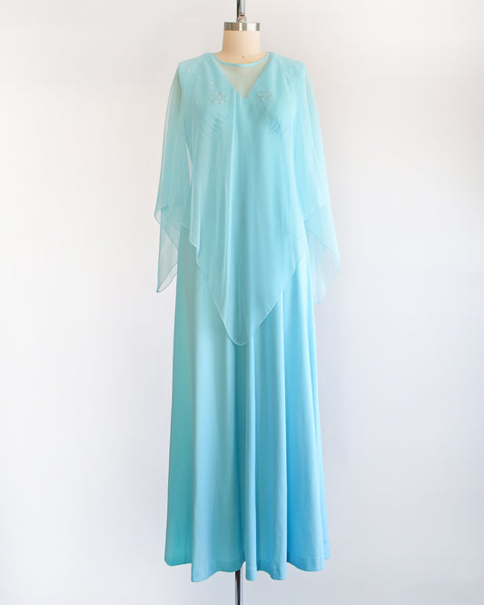 A vintage 70s blue maxi dress with matching semi-sheer blue cape overlay. The dress is on a dress form.