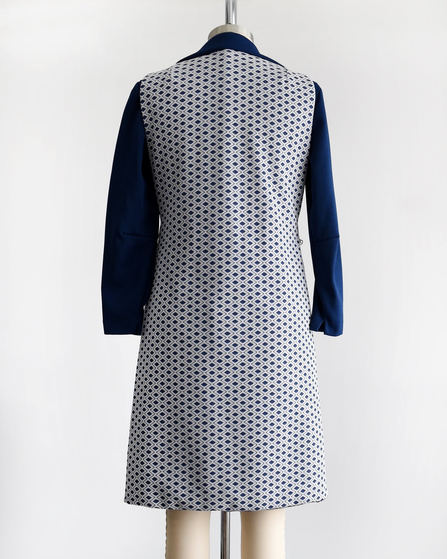 Back view of a vintage 60s/70s two piece dress set that features a long sleeve navy dress with a white and navy diamond print long vest. Dress is modeled on a dress form.