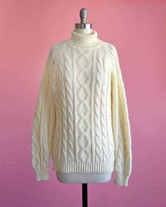 front view of a vintage 70s cream cable knit turtleneck sweater on a dress form set against a pink background