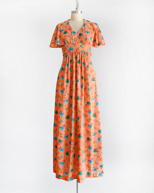 A vintage 70s orange maxi dress with a blue, green, and brown floral print, along with flutter sleeves, a v-neckline, and empire waist. The dress is modeled on a dress form.