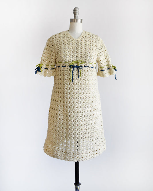 A vintage 70s crochet mini dress with a green and blue woven stripe on the front that ties into a bow, along with matching stripes and bows on the sleeves. The dress is on a dress form.