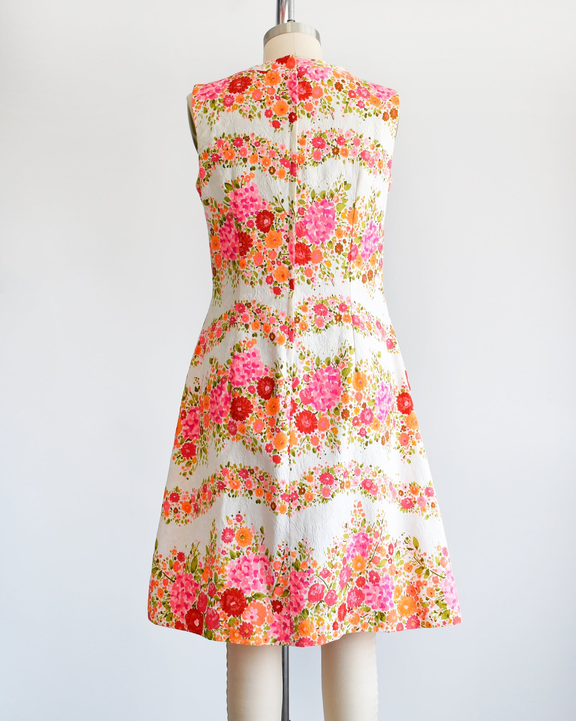 Back view of a vintage 60s/70s white dress that has a vibrant pink, orange, and green floral print in wavy horizontal stripes going down the dress. There is a zipper up the back. The dress is on a dress form.