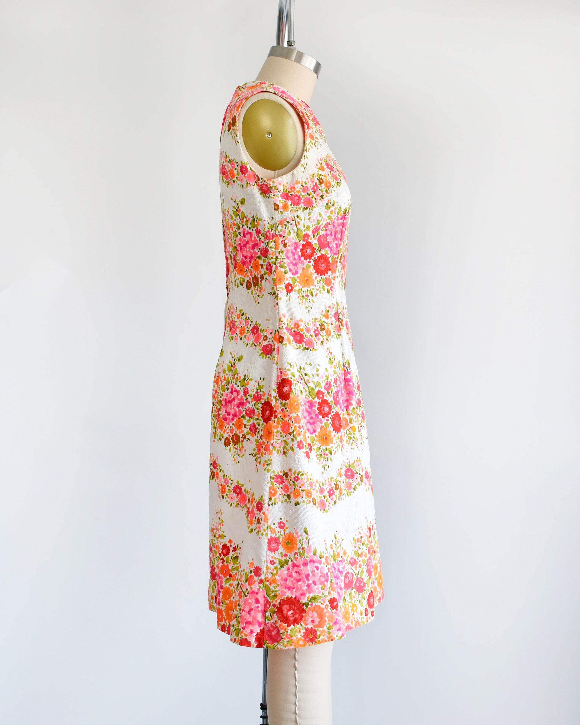 Side view of a vintage 60s/70s white dress that has a vibrant pink, orange, and green floral print in wavy horizontal stripes going down the dress. The dress is on a dress form.