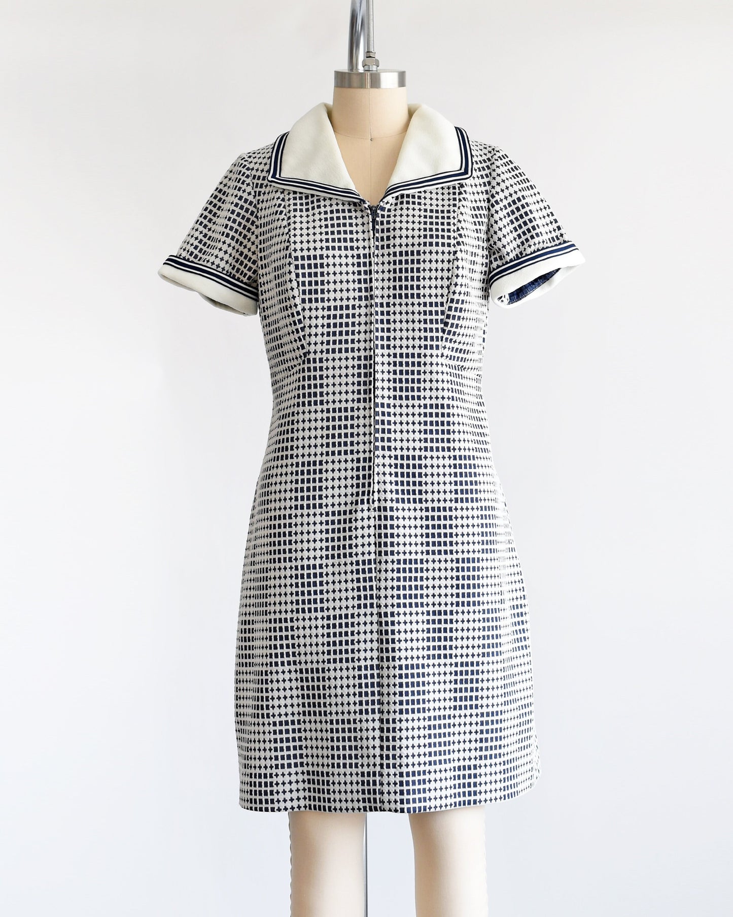 A vintage 70s mod dress that has a blue and white geometric print, white wide collar with navy trim, zip up front, and matching white and navy trim on the short sleeves. The dress is on a dress form.