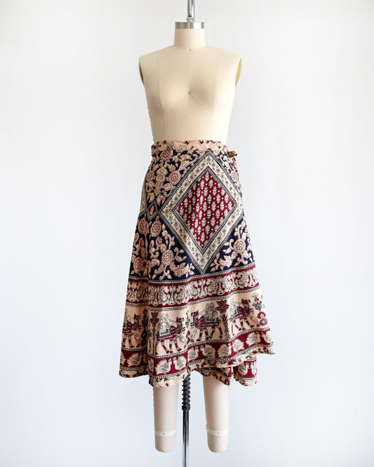 A vintage 1970s cotton wrap skirt that is beige with a large burgundy and black batik block floral print that covers the entire skirt, along border prints of people riding camels and a bird print. Skirt is on a dress form.