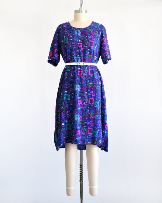 A vintage floral dress that has a vibrant purple, blue, pink, teal, green, and white psychedelic floral print. The dress has short sleeves, scoop neckline, and is modeled with a white belt on a dress form.