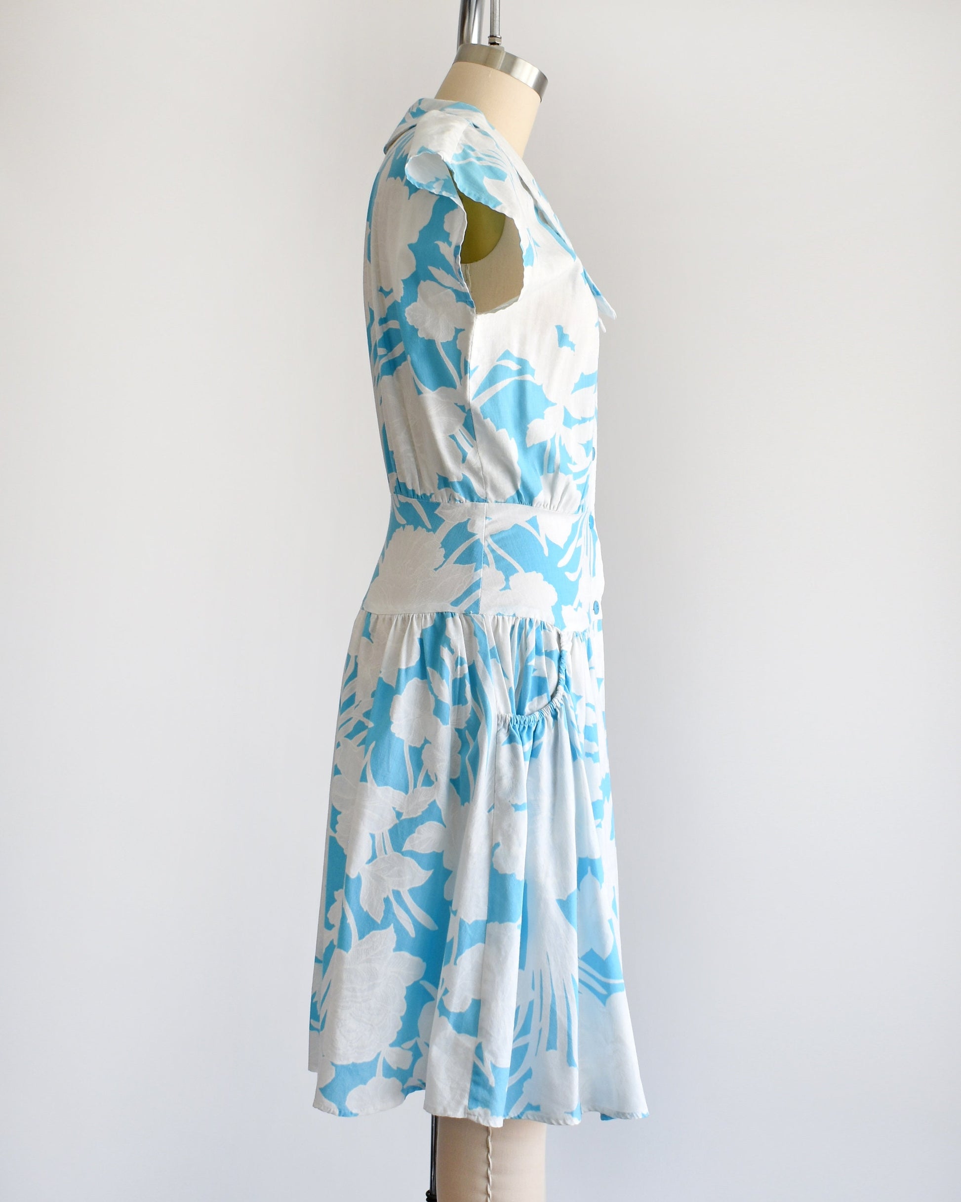 A vintage 80s floral dress that is light blue with white floral print. Cap sleeves. Wide waistband and drop waist skirt.