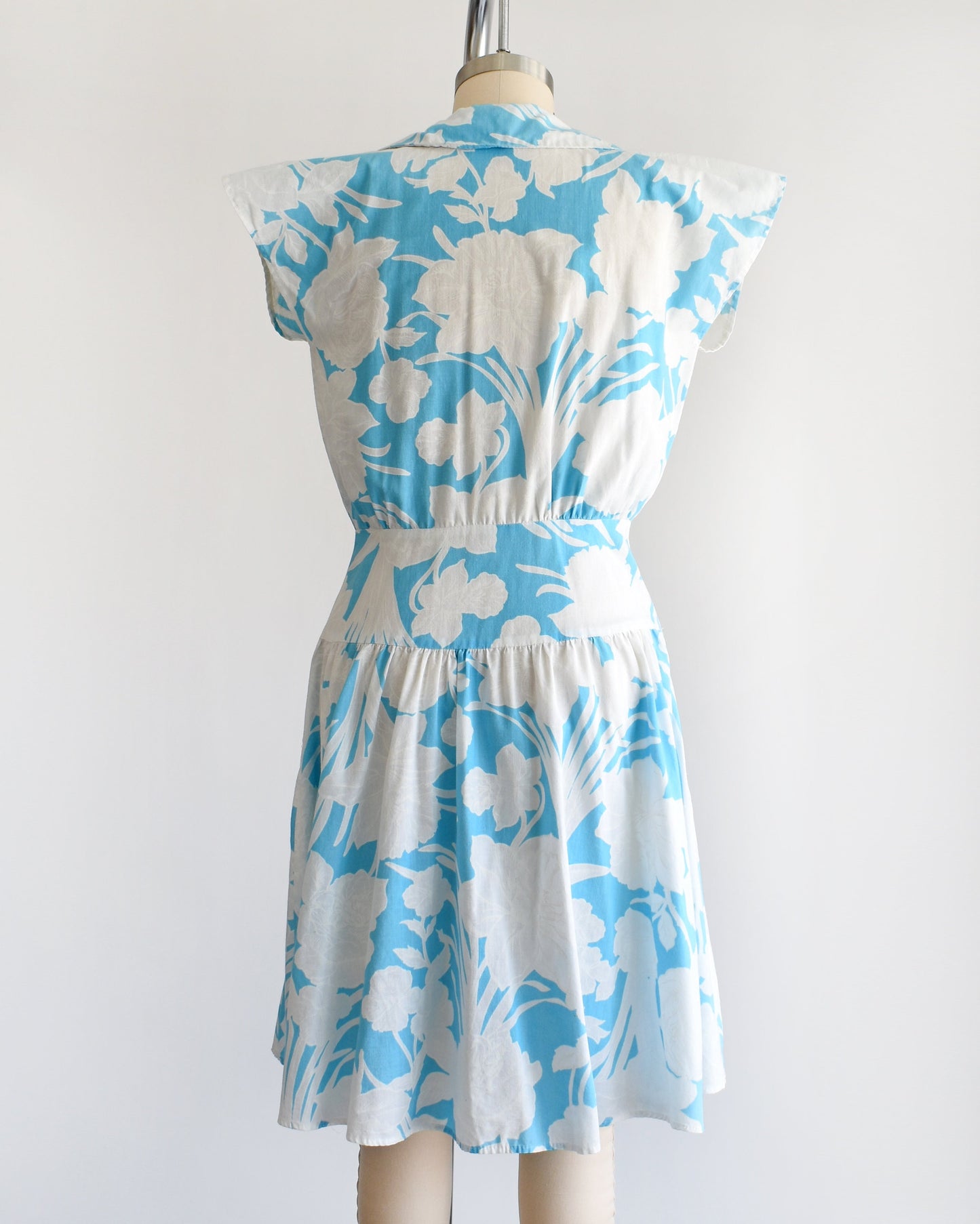 Back view of a vintage 80s floral dress that is light blue with white floral print. Cap sleeves. Wide waistband and drop waist skirt.