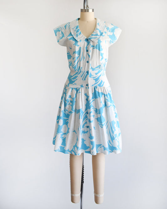 A vintage 80s floral dress that is light blue with white floral print. Peter Pan style collar with blue buttons down the front. Cap sleeves. Wide waistband and drop waist skirt.