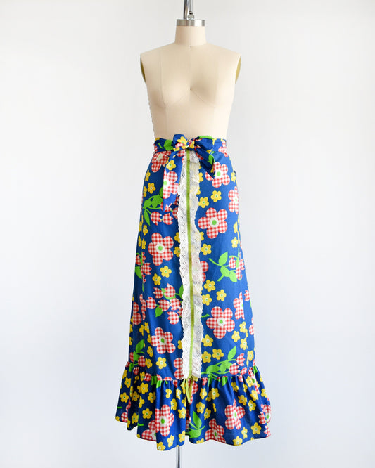 A vintage 70s maxi skirt that is blue with a red and white gingham floral print, along with yellow and green flowers with green leaves and stems. White eyelet trim down the middle of the skirt with a velvet green ribbon down the middle. Matching belt