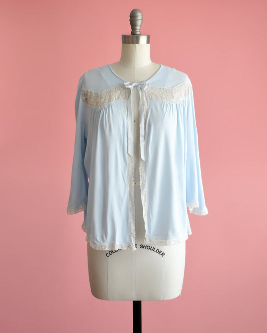 Front view of a vintage 60s light blue bed jacket with cream lace trim around the front, down the front, around the hem, and cuffs. A ribbon tie is at the center of the collar. The garment is modeled on a dress form.
